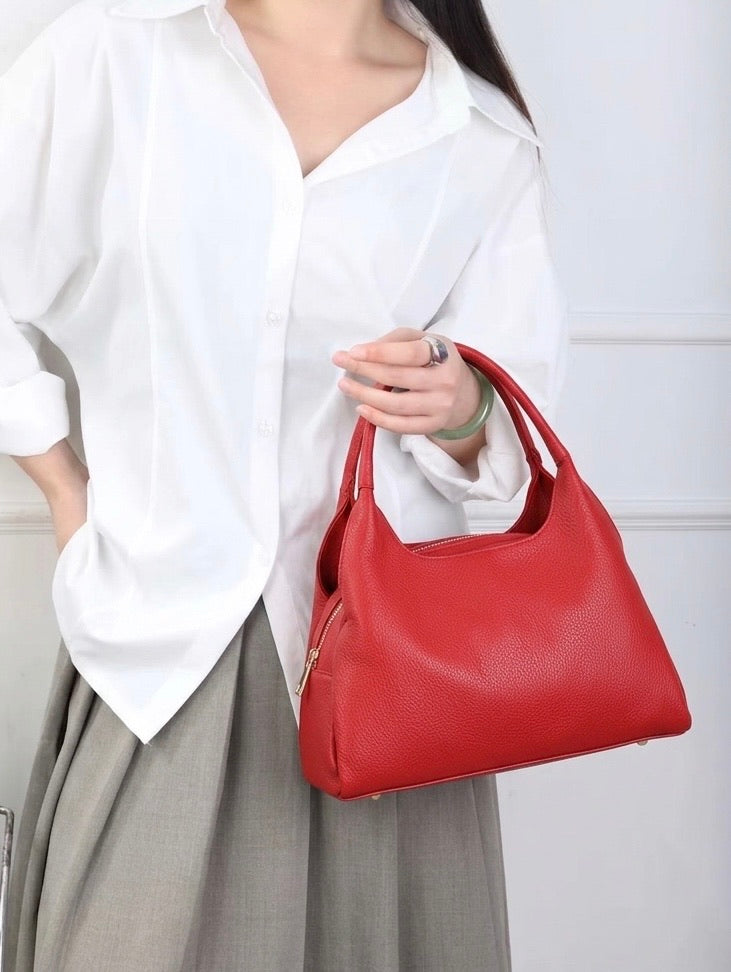 A woman wearing a white button-down shirt and gray skirt holds a red pebbled leather shoulder bag with gold-tone hardware.